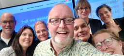 Selfie van een bijeenkomst. 8 deelnemers kijken in de camera terwijl ze voor een digitaal bord staan.