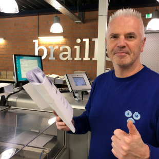 Een werknemer staat in de ruimte bij de brailleprinter. Hij houdt een brailleboek vast en steekt zijn duim op.